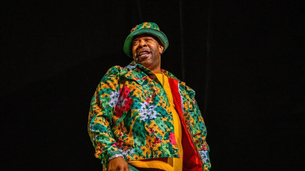 Busta Rhymes on stage in a patterned jacket and bucket hat against a dark backdrop