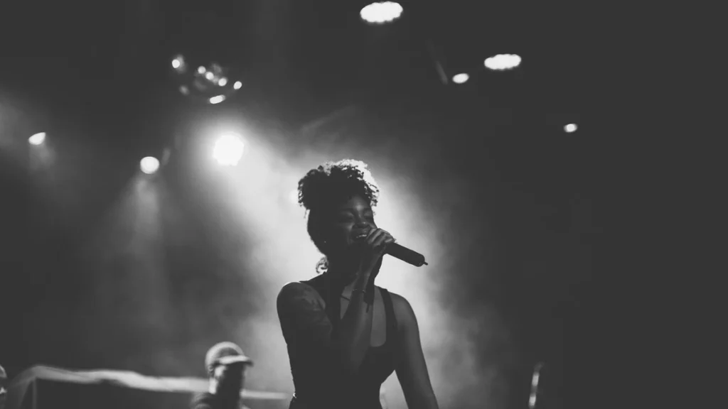 Ari Lennox singing into a microphone under stage haze in black and white