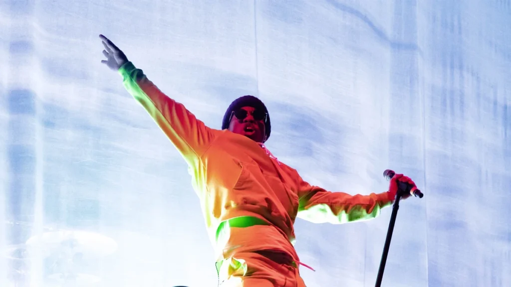 Anderson .Paak performing on stage in a bright orange outfit with arms outstretched