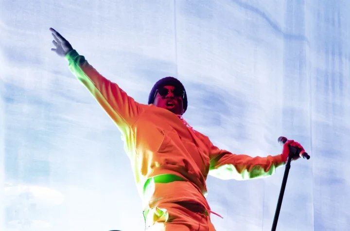 Anderson .Paak performing on stage in a bright orange outfit with arms outstretched