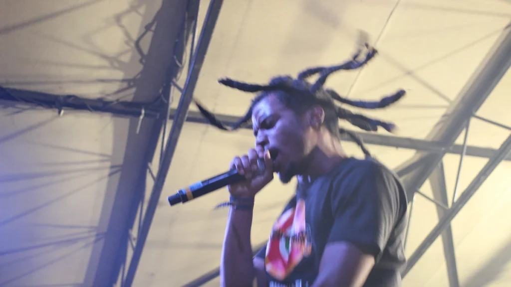 Denzel Curry rapping into a microphone on stage with his locs mid-motion
