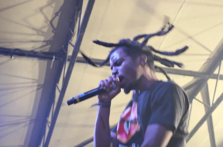 Denzel Curry rapping into a microphone on stage with his locs mid-motion